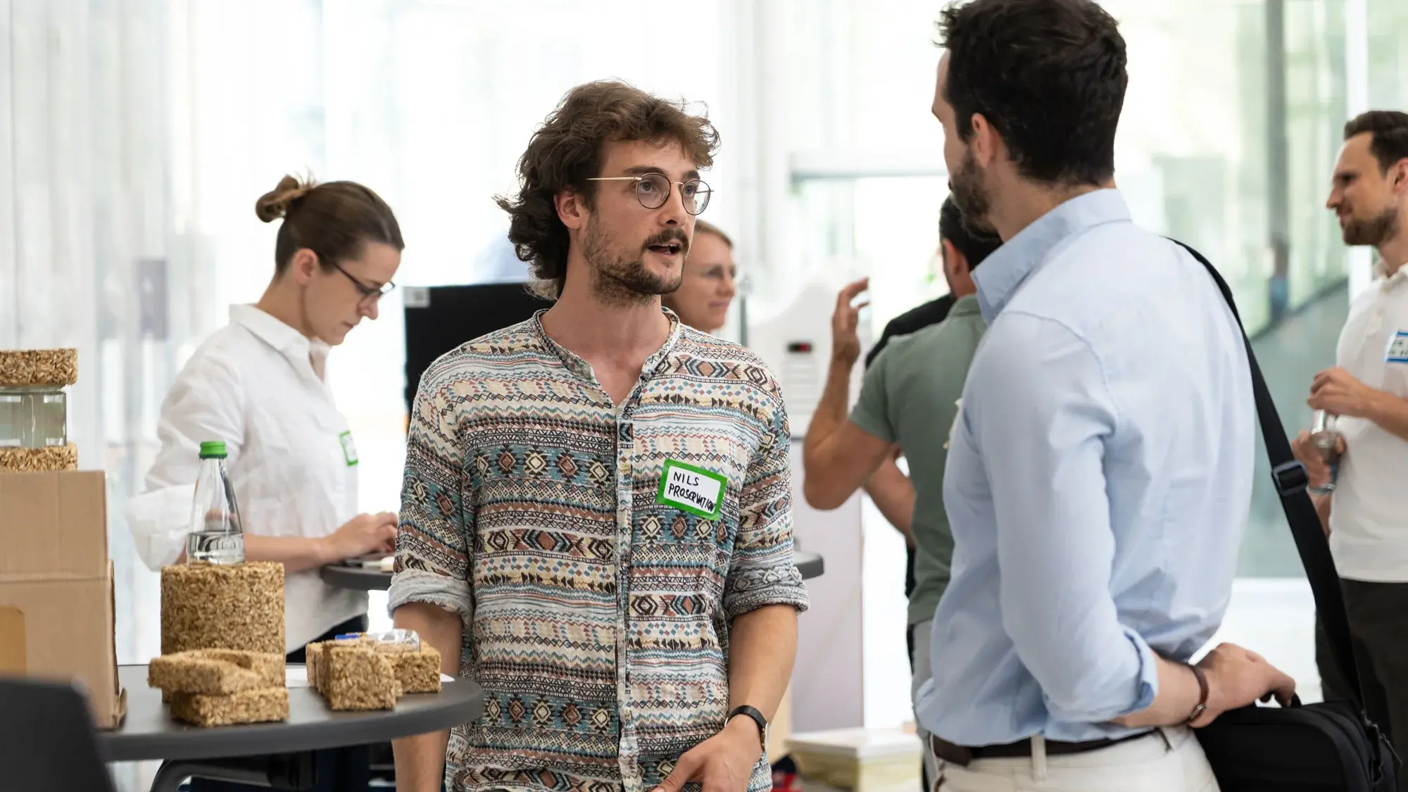 Zwei Personen diskutieren an einem Messestand. Auf dem Stand stehen Verpackungsmaterialien eines Startups