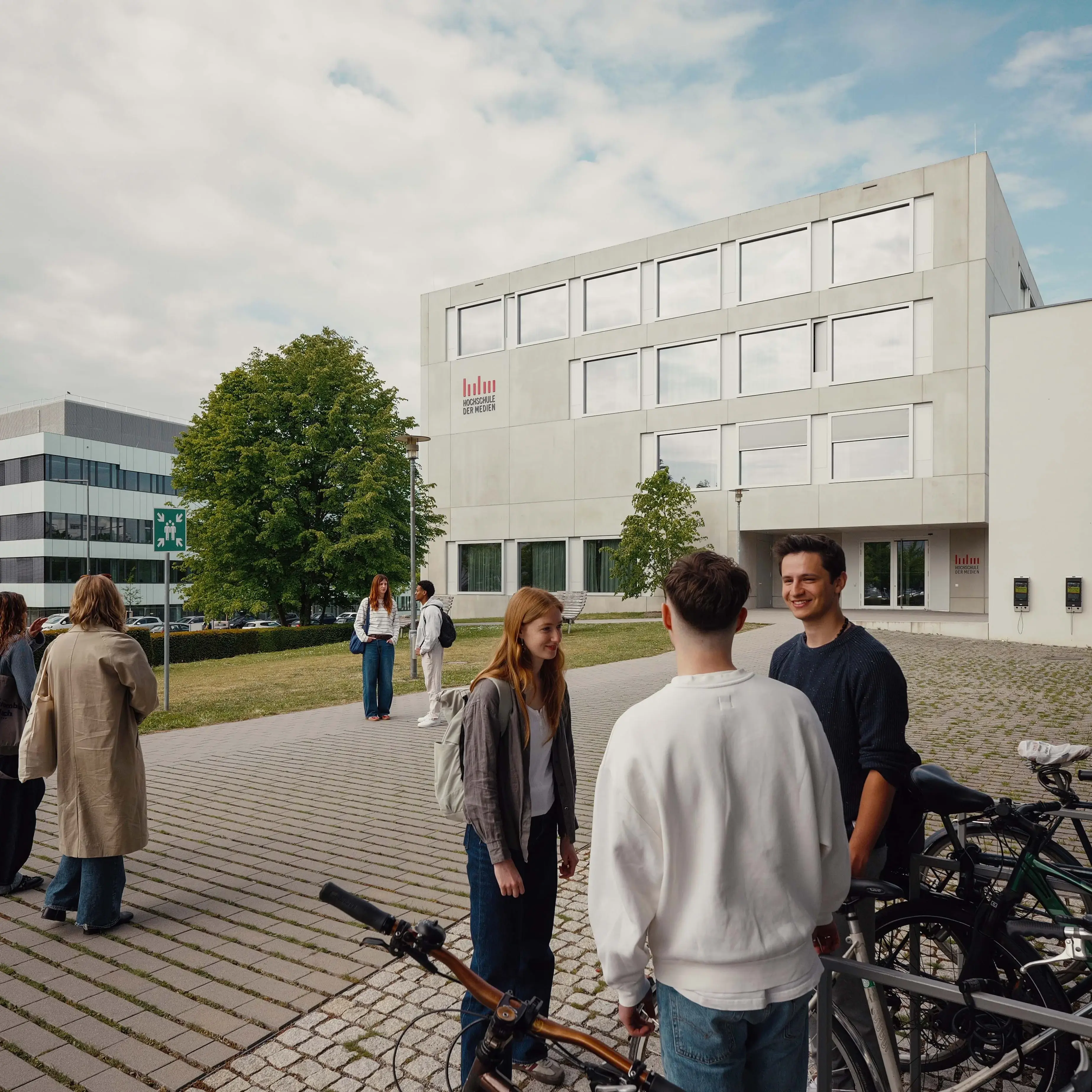 Studierende unterhalten sich bei den Fahrradständern vor einem Nebengebäude der Hochschule der Medien.